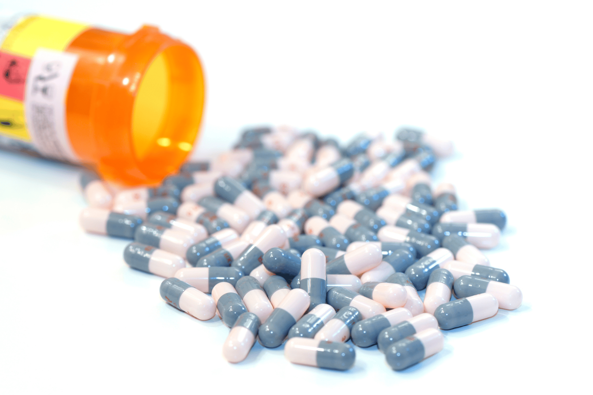 A prescription bottle tipped over with many grey-and-pink capsules spilled across a white surface.