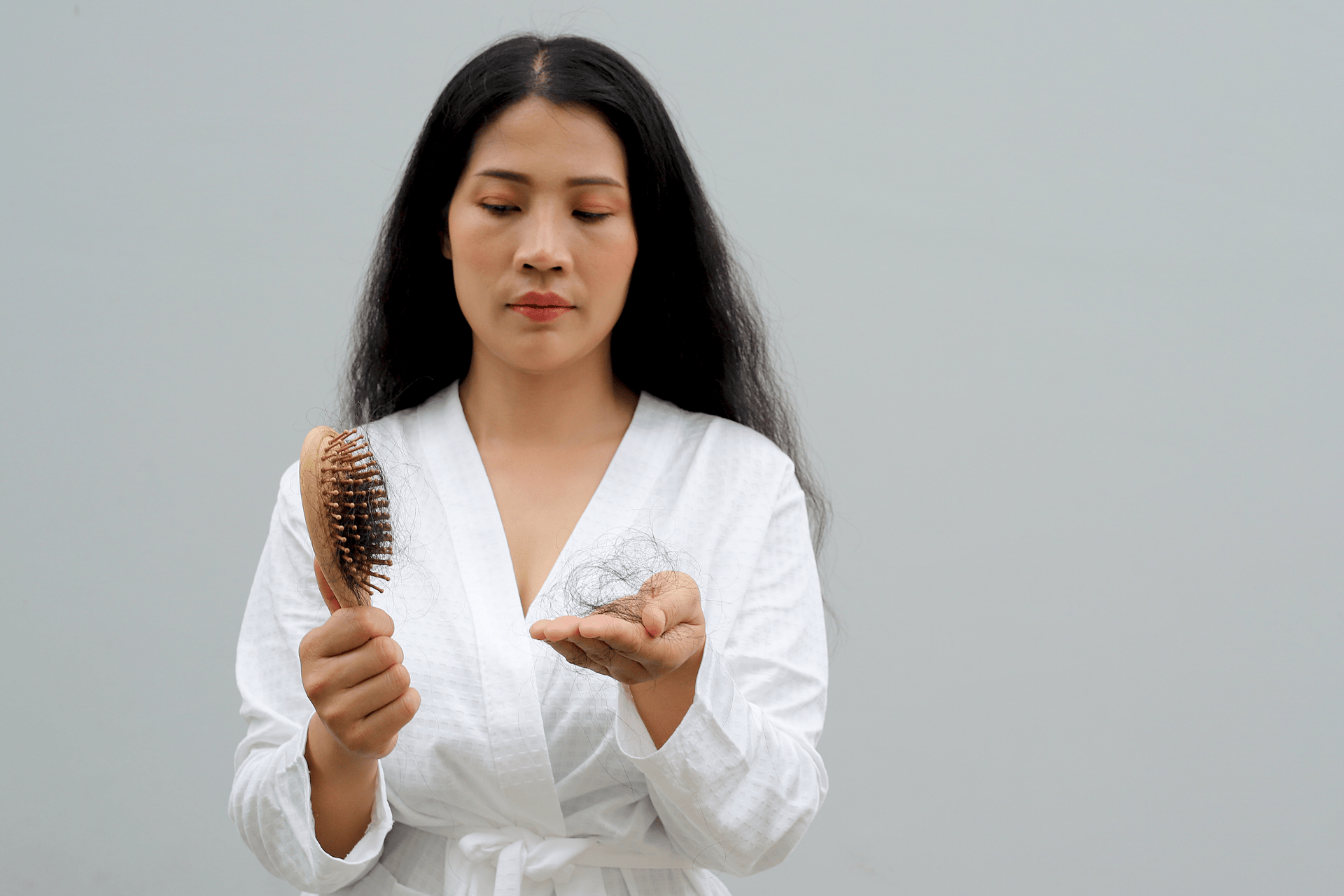 Woman in a white robe looking at a hairbrush and holding a clump of fallen hair in her hand, appearing concerned about hair loss.