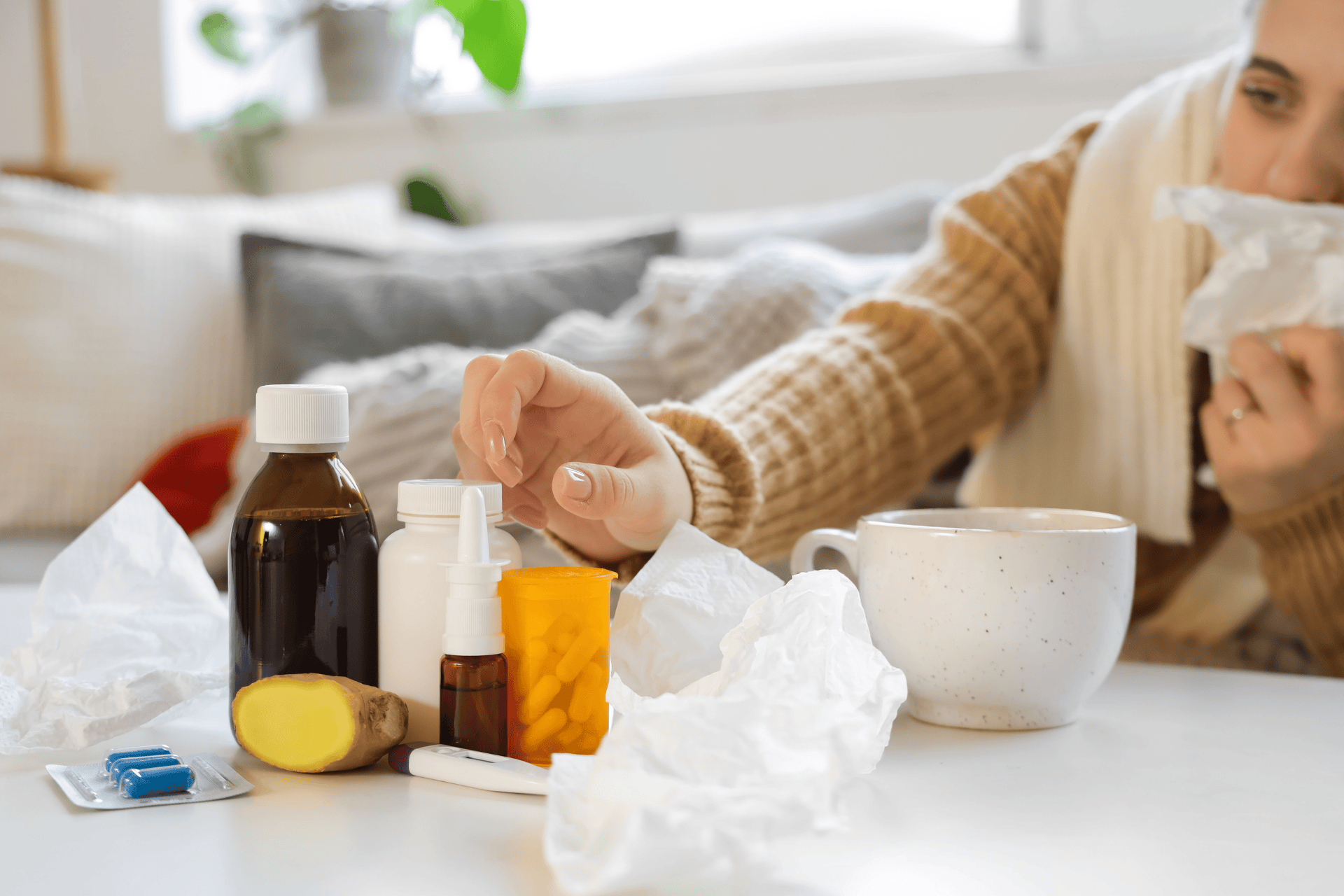 Sick person sitting at a table surrounded by cold and flu items, including tissues, medicine bottles, pills, a nasal spray, ginger, and a mug, while holding a tissue to their nose.