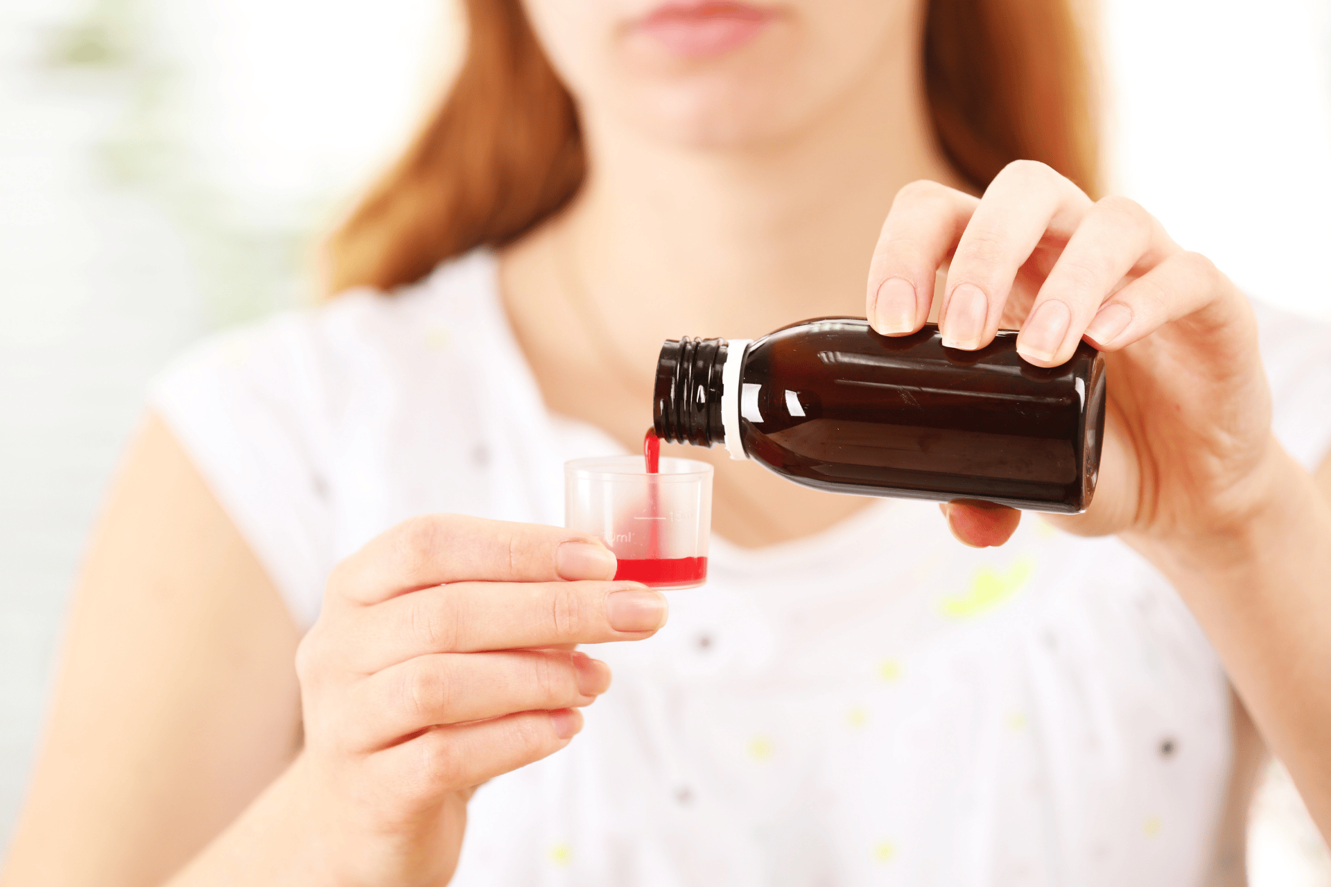 Woman pouring red cough medicine from a dark bottle into a small measuring cup.