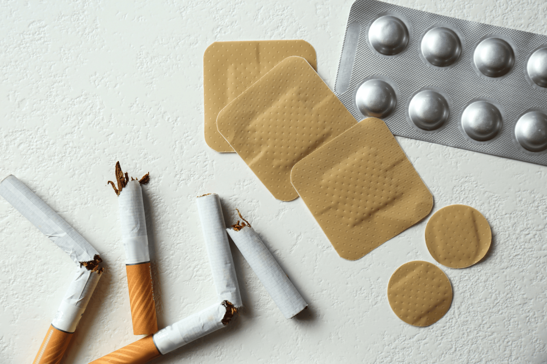 Broken cigarettes, nicotine patches, and a blister pack of pills arranged on a light textured surface, representing smoking cessation tools.