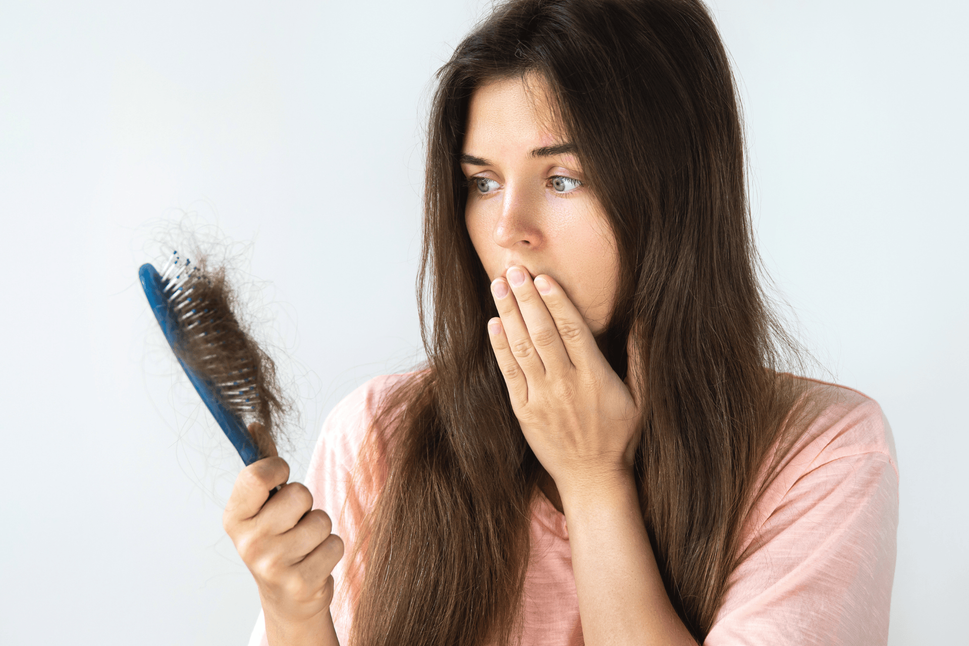 Woman looking shocked while holding a hairbrush full of shed hair, covering her mouth with her hand.