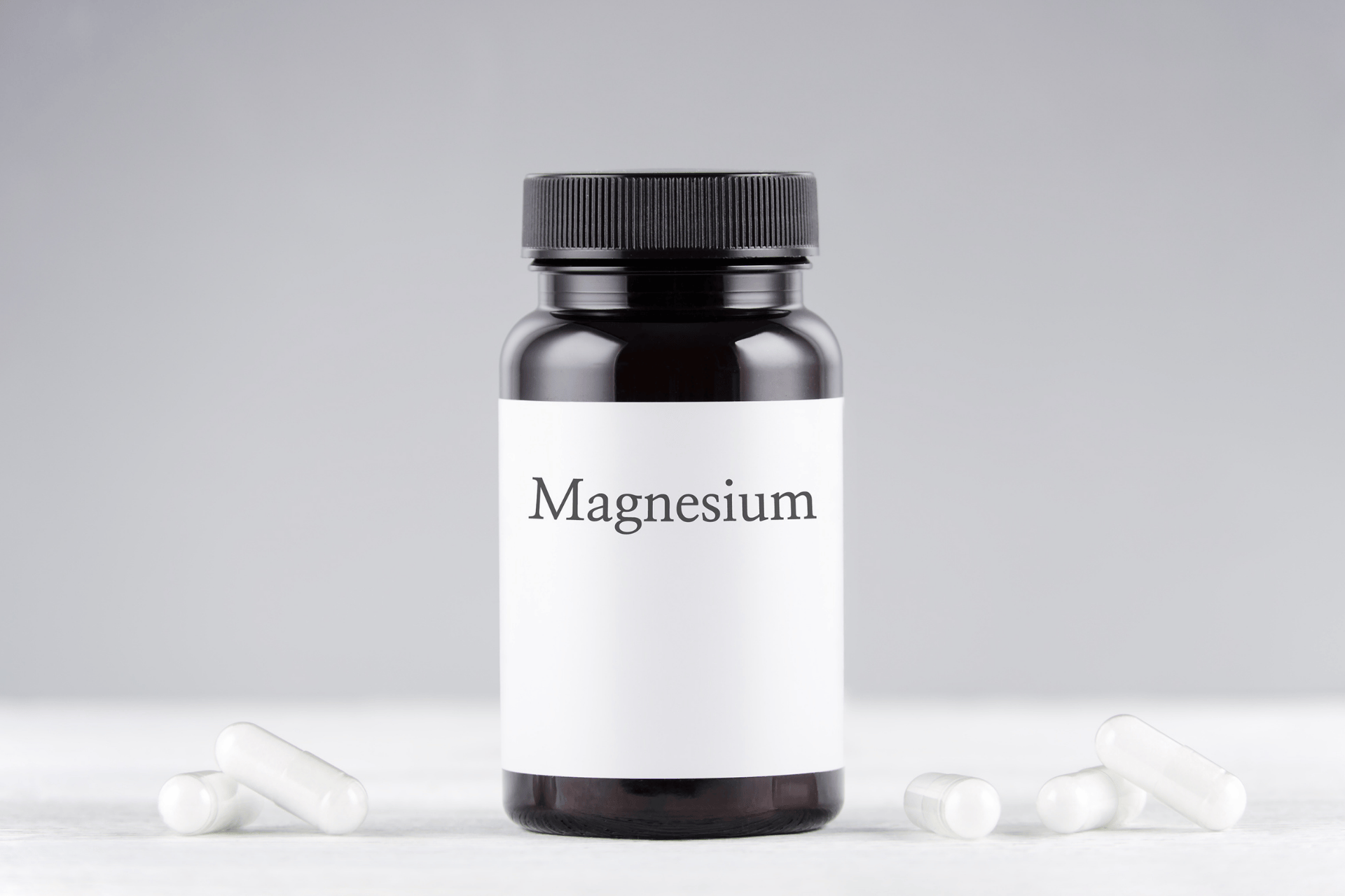 A black supplement bottle labeled “Magnesium” surrounded by several white capsules on a light surface.