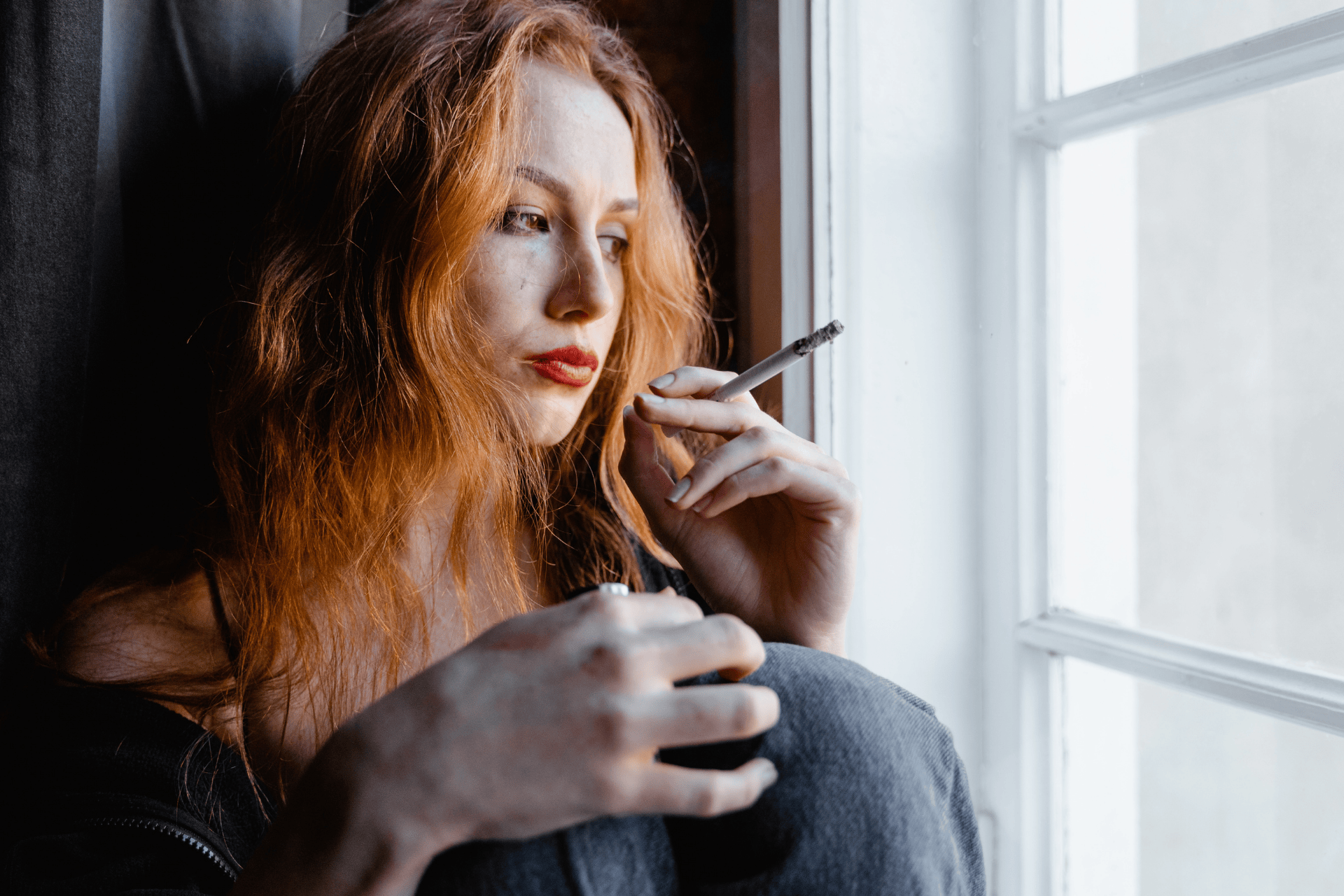 A woman with long red hair sits by a window, holding a cigarette and looking contemplative or distressed.