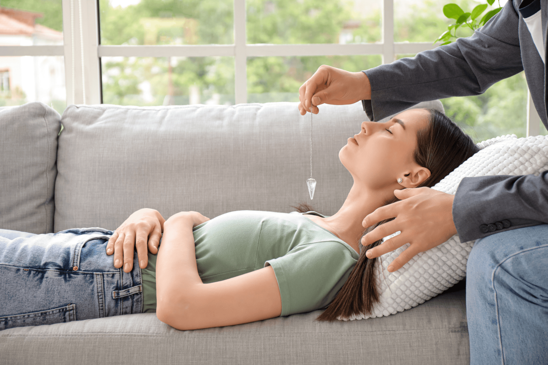 A woman lying on a couch with eyes closed while a practitioner holds a pendulum above her, suggesting hypnosis or relaxation therapy.