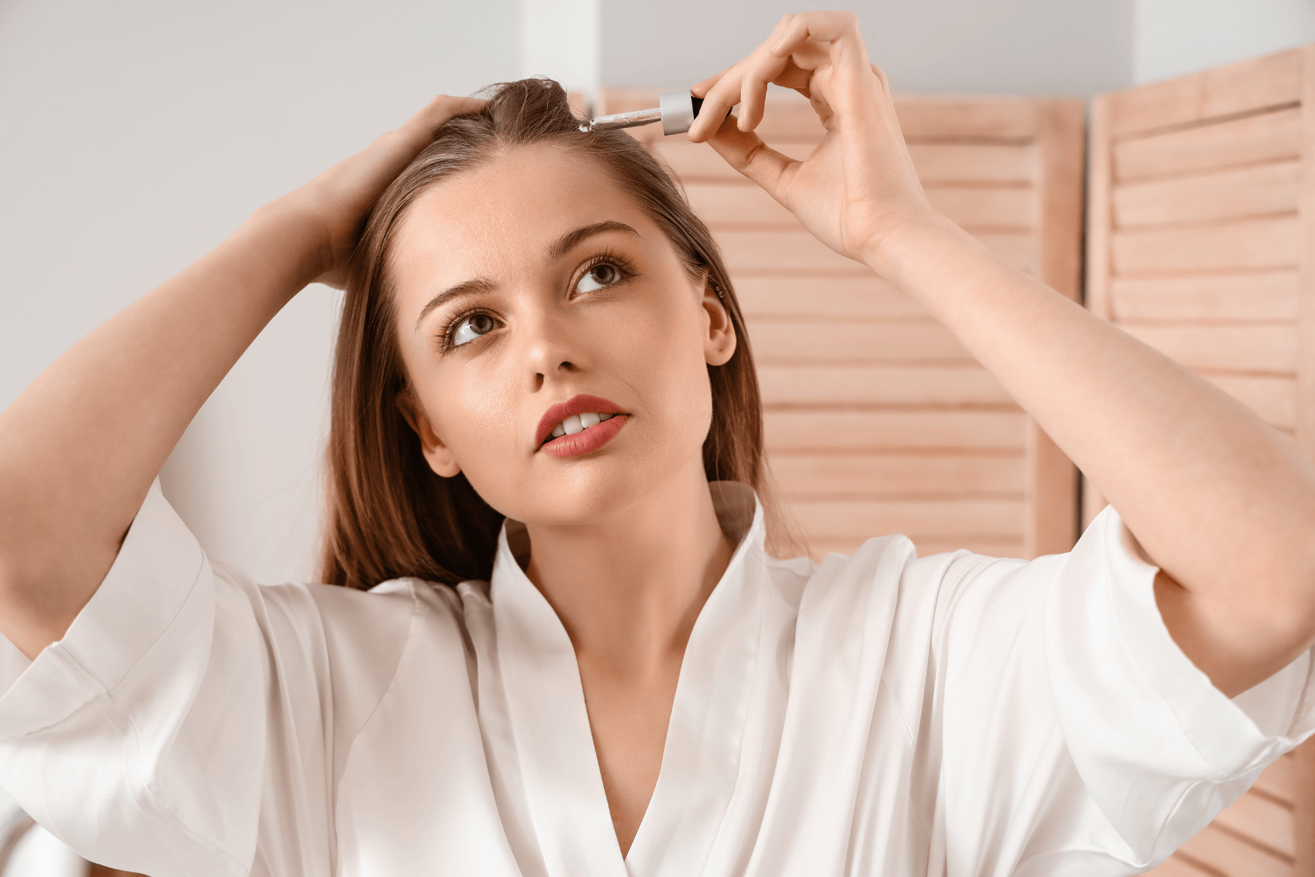 A woman applying a hair serum to her scalp using a dropper.