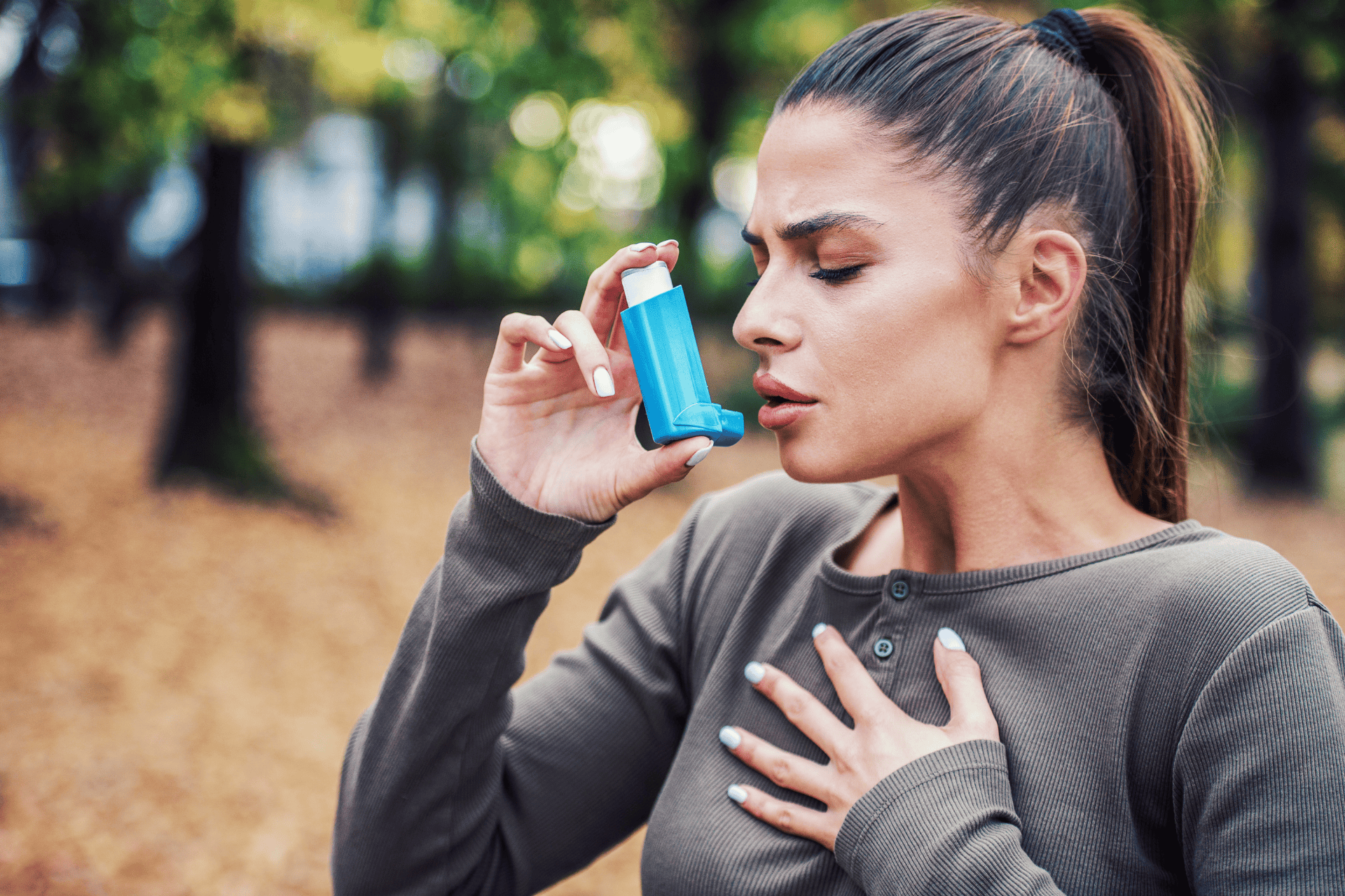A woman using an inhaler outdoors while holding her chest.