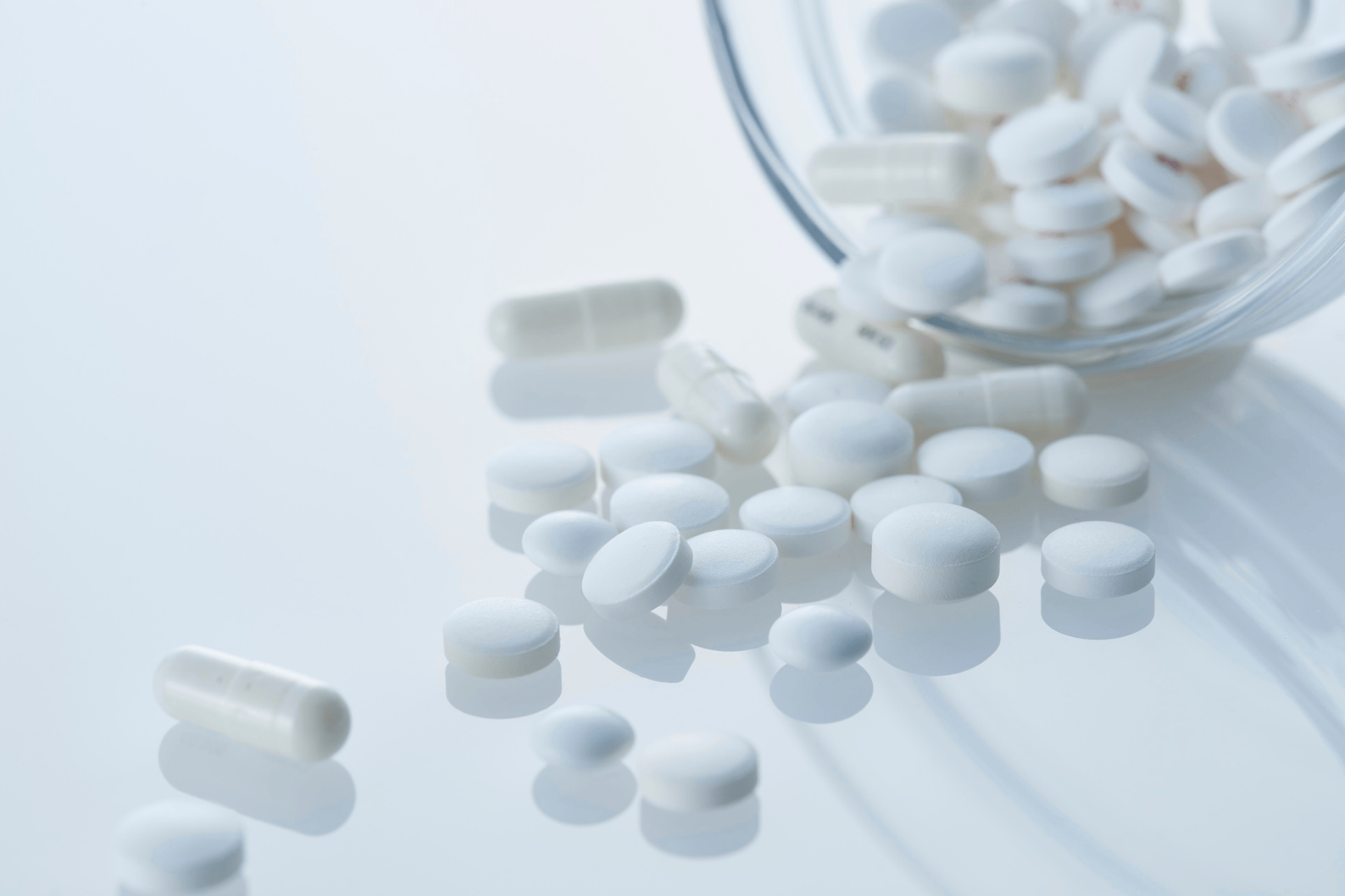 White pills and capsules spilled from a clear container on a white surface.