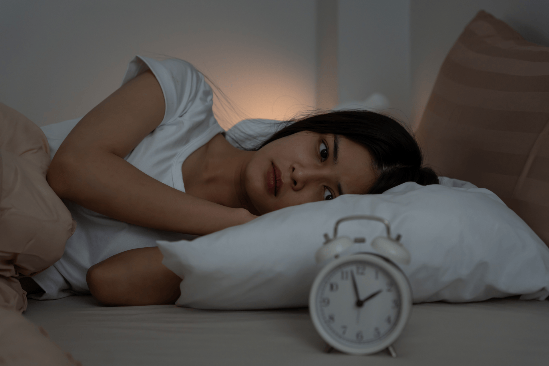Woman lying awake in bed at night, looking tired, with an alarm clock in the foreground.