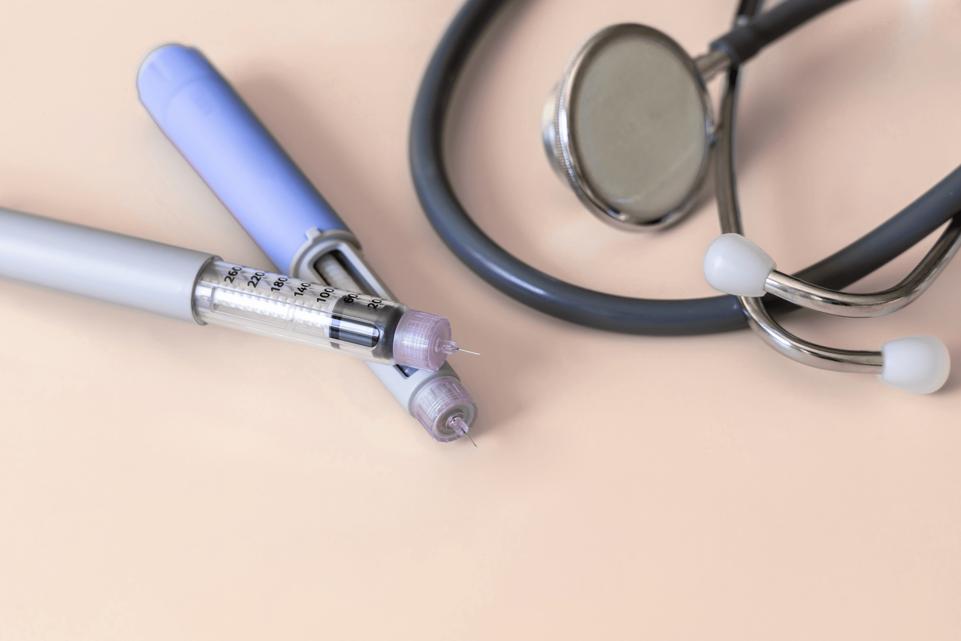A close-up photo of two medical syringes with needles resting on a light beige surface beside a black stethoscope, suggesting a healthcare or medical setting.