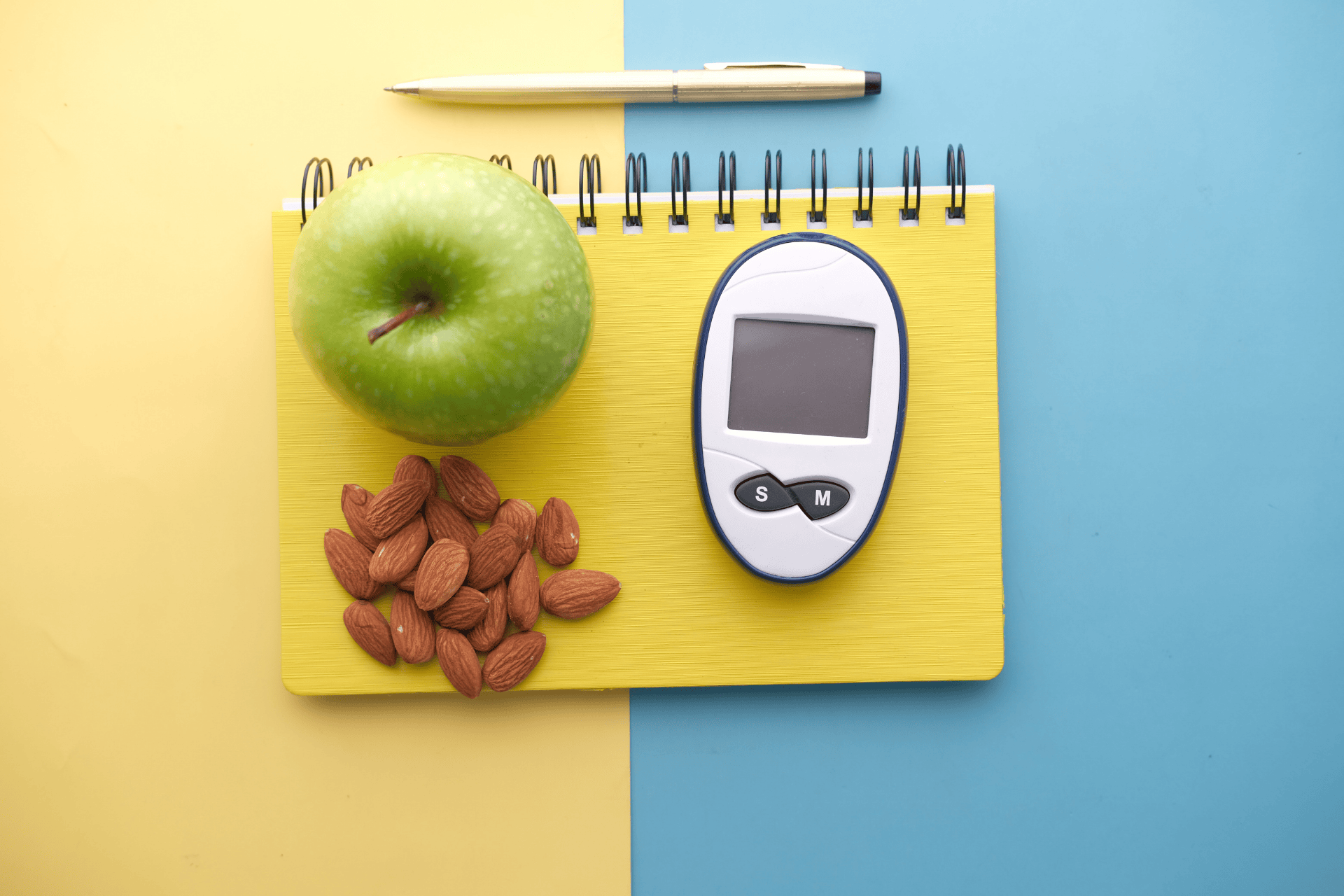 A glucose meter beside an apple and almonds on a notebook, symbolizing healthy diabetes management.