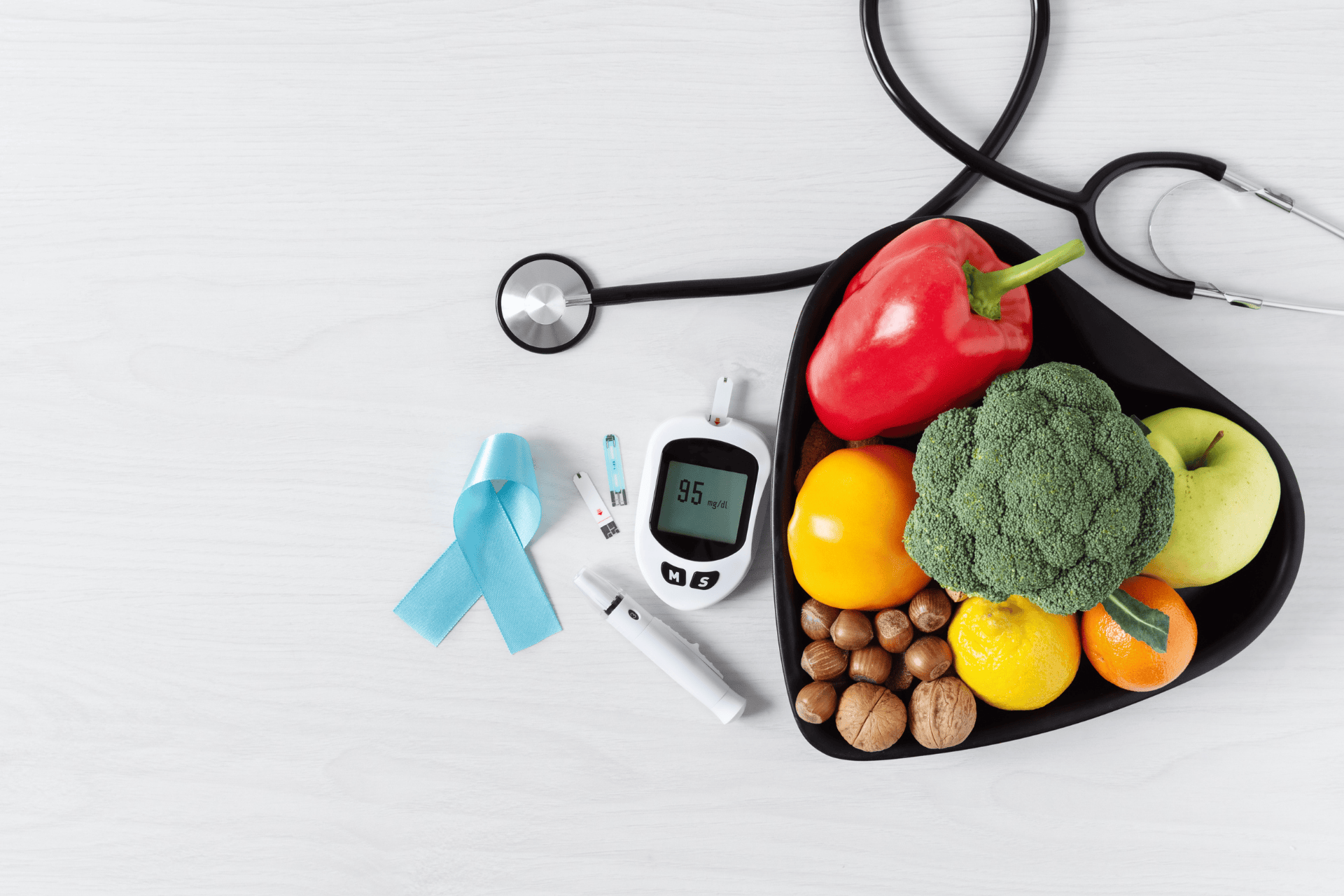 A heart-shaped bowl of fruits and vegetables with a glucose meter, stethoscope, and diabetes awareness ribbon.