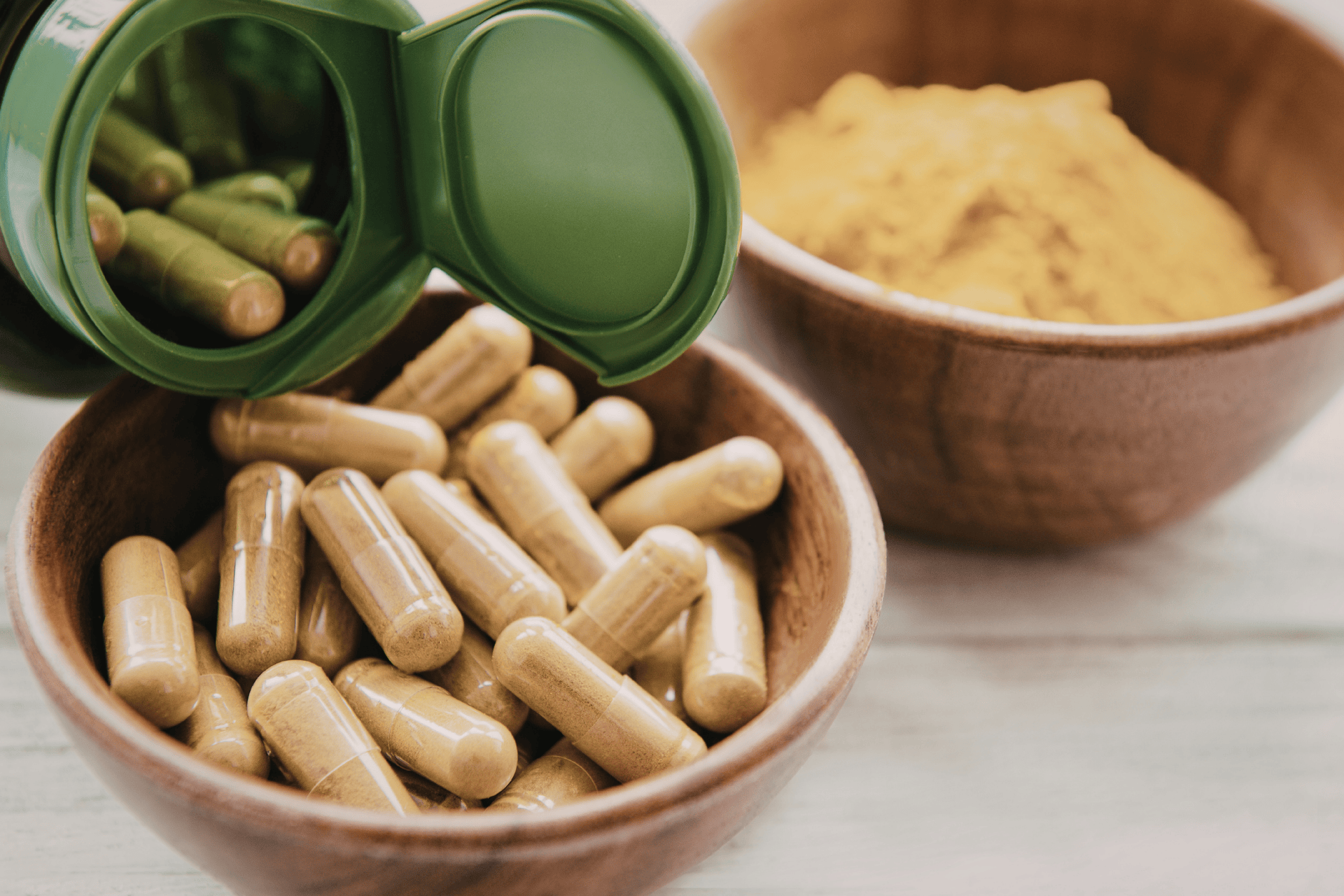 Herbal supplement capsules poured from a green container into a wooden bowl, with powdered herbs in the background.
