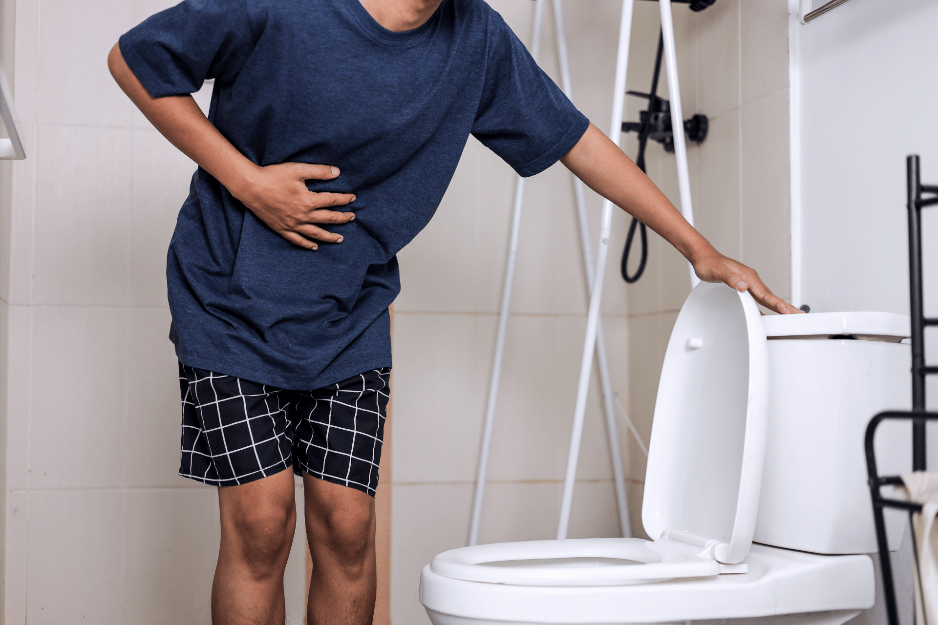 Person standing in a bathroom holding their stomach and leaning toward a toilet, suggesting diarrhea or abdominal discomfort.