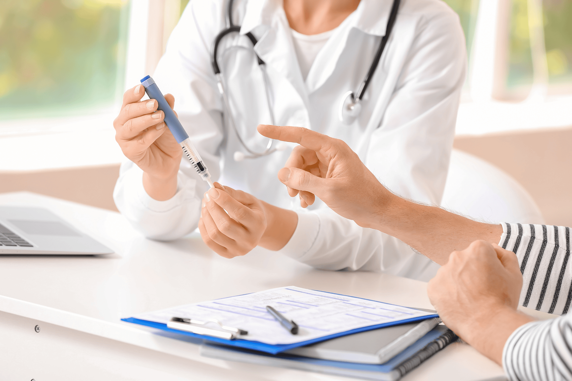 A healthcare professional in a white coat holding an insulin pen while explaining it to a patient, who points at the device during a consultation at a desk with paperwork.