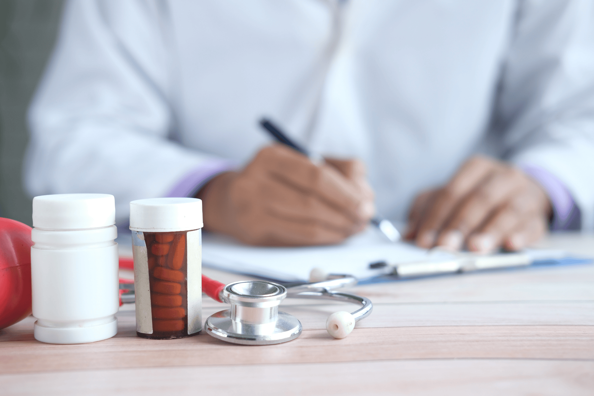A healthcare professional writing a prescription, with medication bottles and a stethoscope in the foreground.