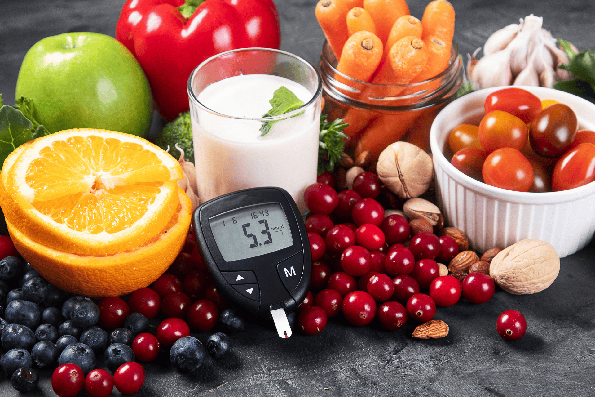 Blood glucose meter surrounded by fruits, vegetables, nuts, and a glass of milk on a dark surface.