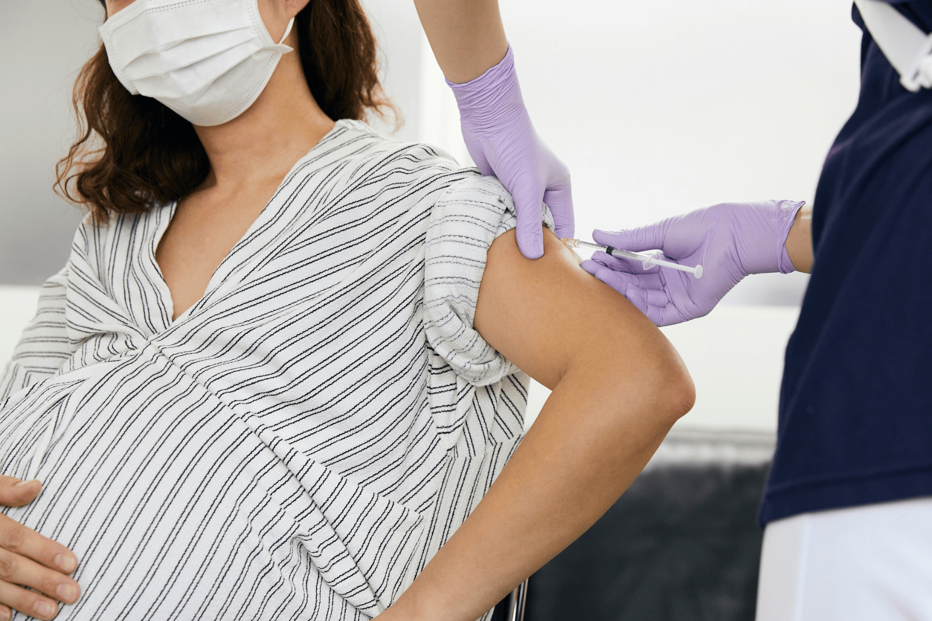 Pregnant woman wearing a mask receiving a flu shot in her upper arm from a healthcare provider.