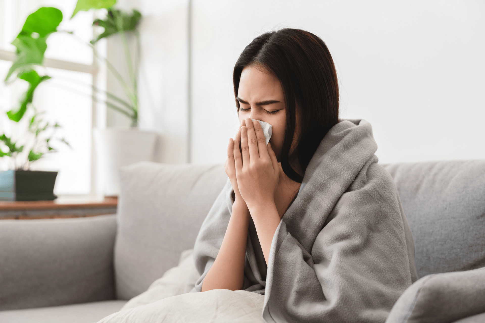 Woman wrapped in a blanket sitting on a couch, blowing her nose with a tissue during flu recovery.