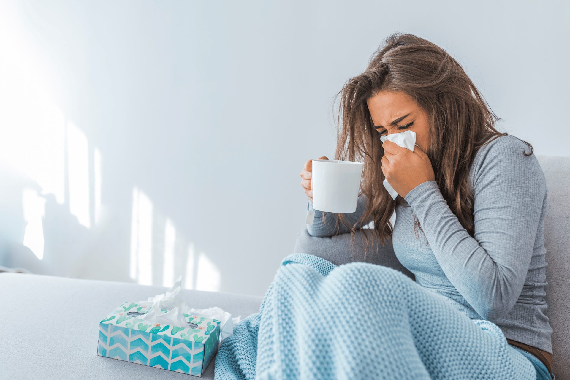 Woman sitting on a couch with a blanket, blowing her nose into a tissue while holding a mug, with a box of tissues beside her, appearing sick or congested.