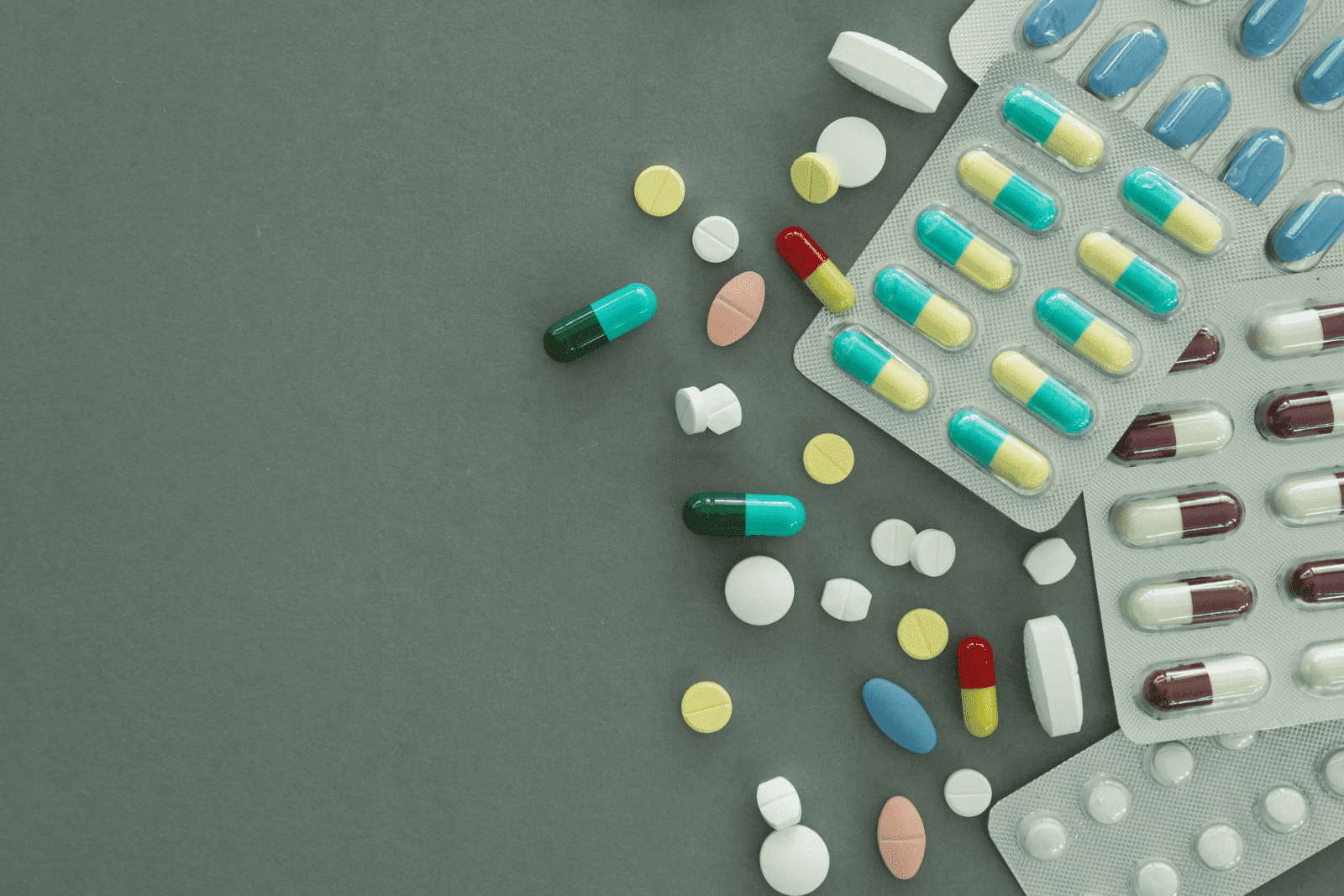 Various pills and capsules scattered next to blister packs on a dark green surface.