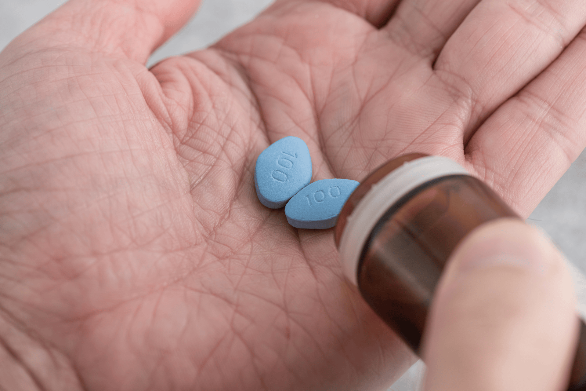 Hand holding blue, oval tablets resembling Viagra being poured from a small pill bottle.