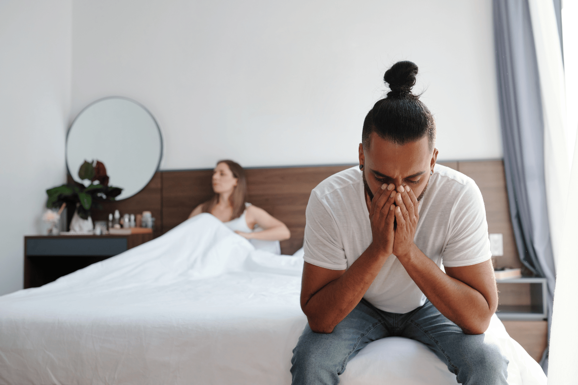A Man sitting on the edge of a bed with his face in his hands, looking distressed, while a woman sits in the background under the covers.