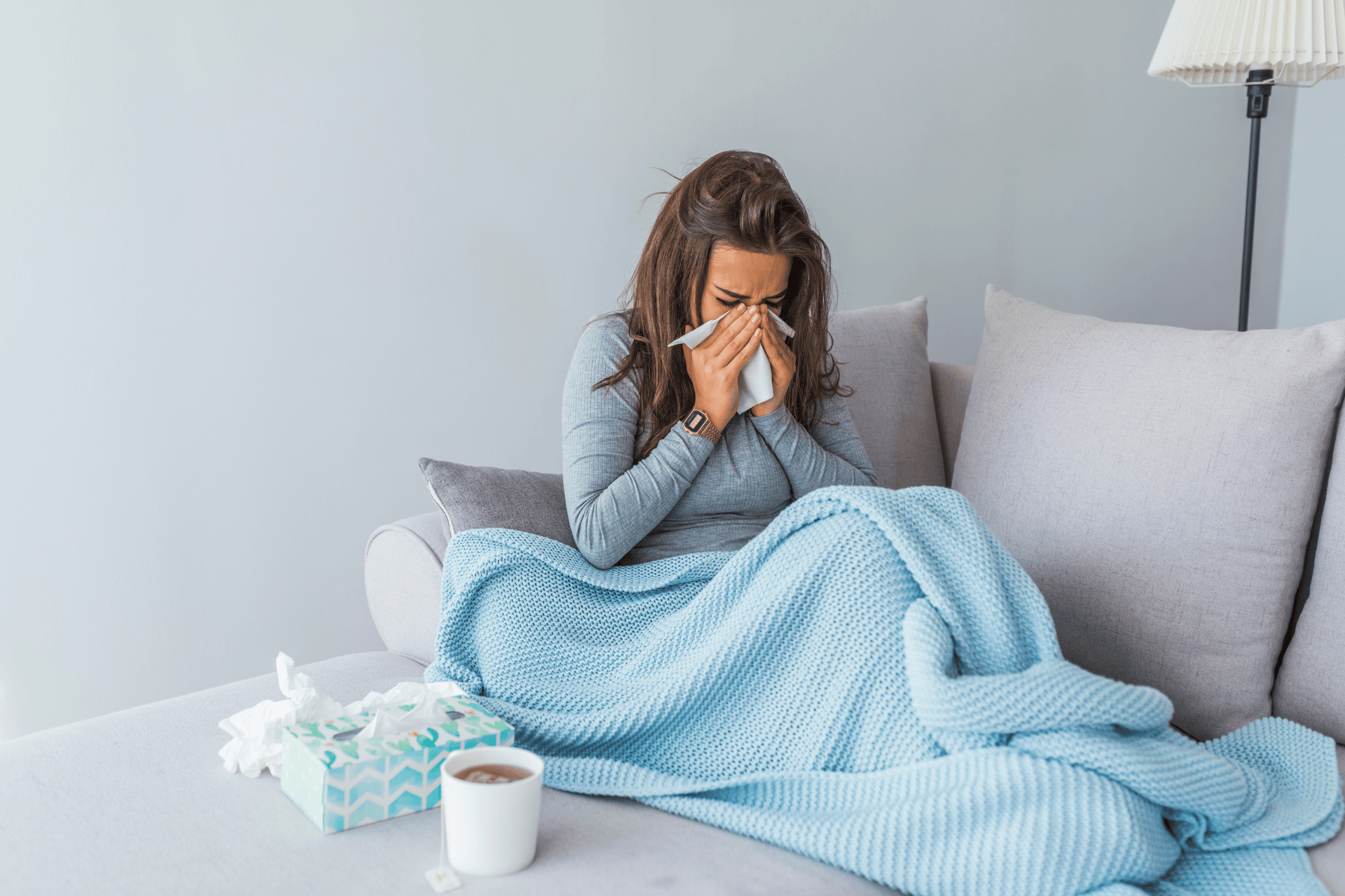 Woman sitting on a couch with a blanket, blowing her nose and appearing sick, with tissues and a hot drink nearby.
