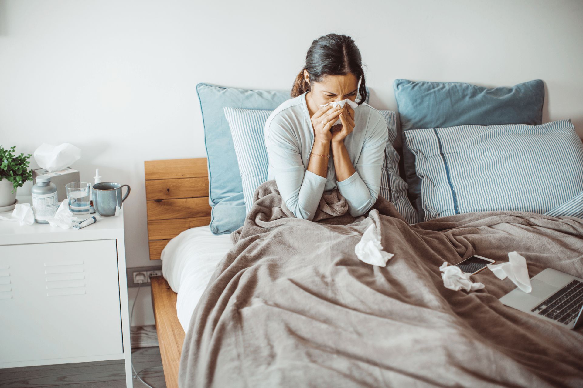 A woman sitting in bed blowing her nose, surrounded by tissues, a blanket, and cold-remedy items on a bedside table