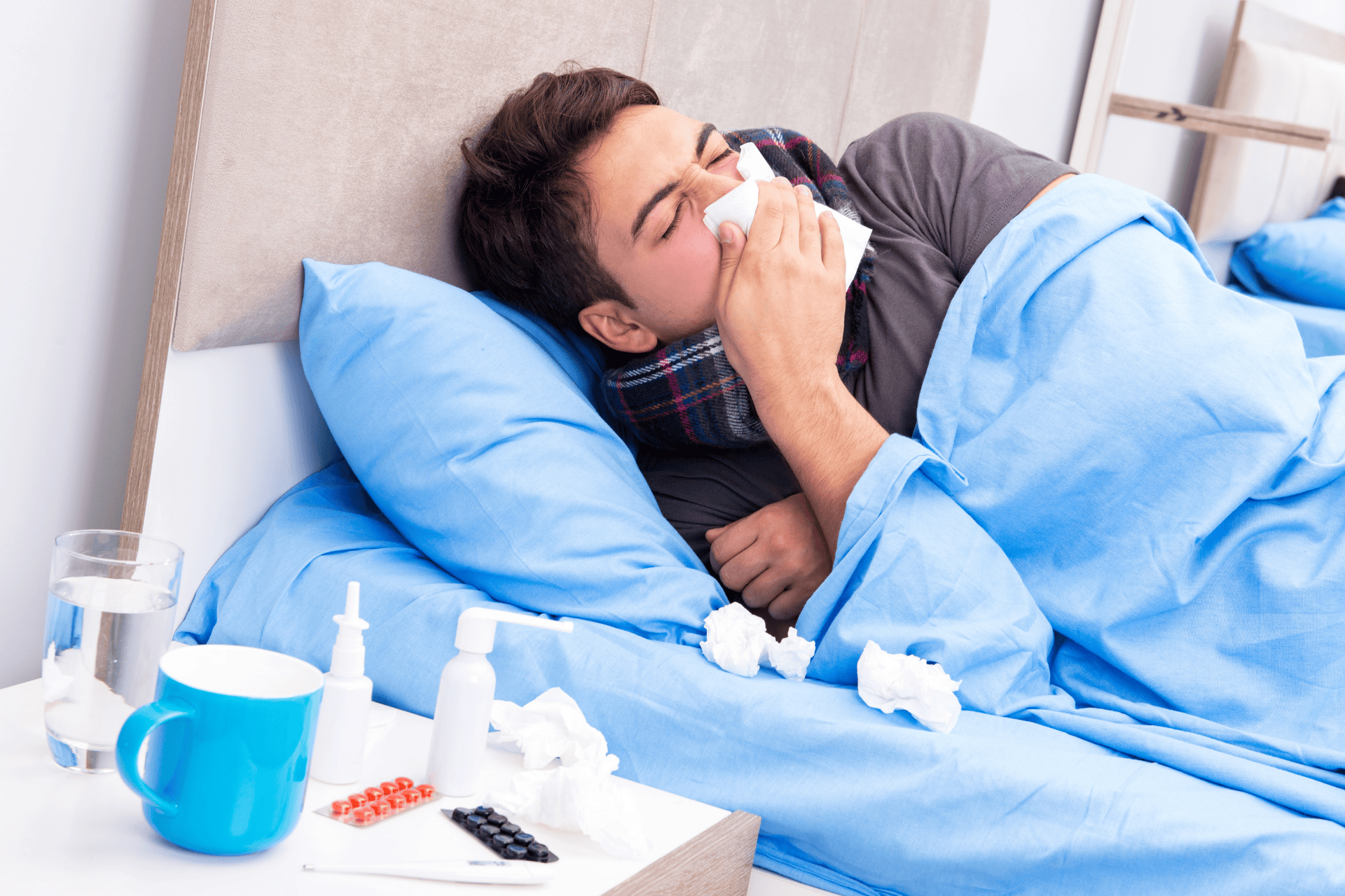 Man lying in bed with a tissue to his nose, surrounded by flu medicine, tissues, and a mug on a bedside table.