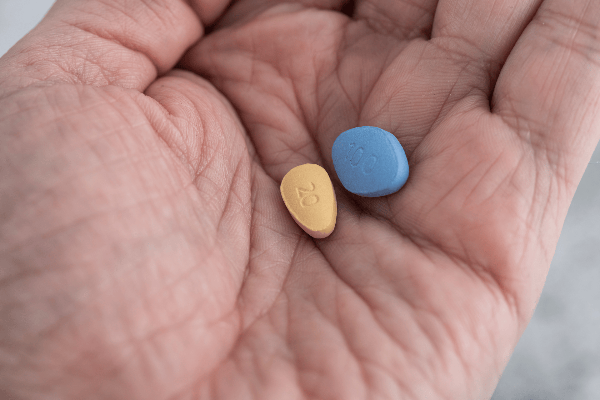 Hand holding a blue 100 mg pill and a yellow 20 mg pill, representing Viagra and a similar erectile dysfunction medication.