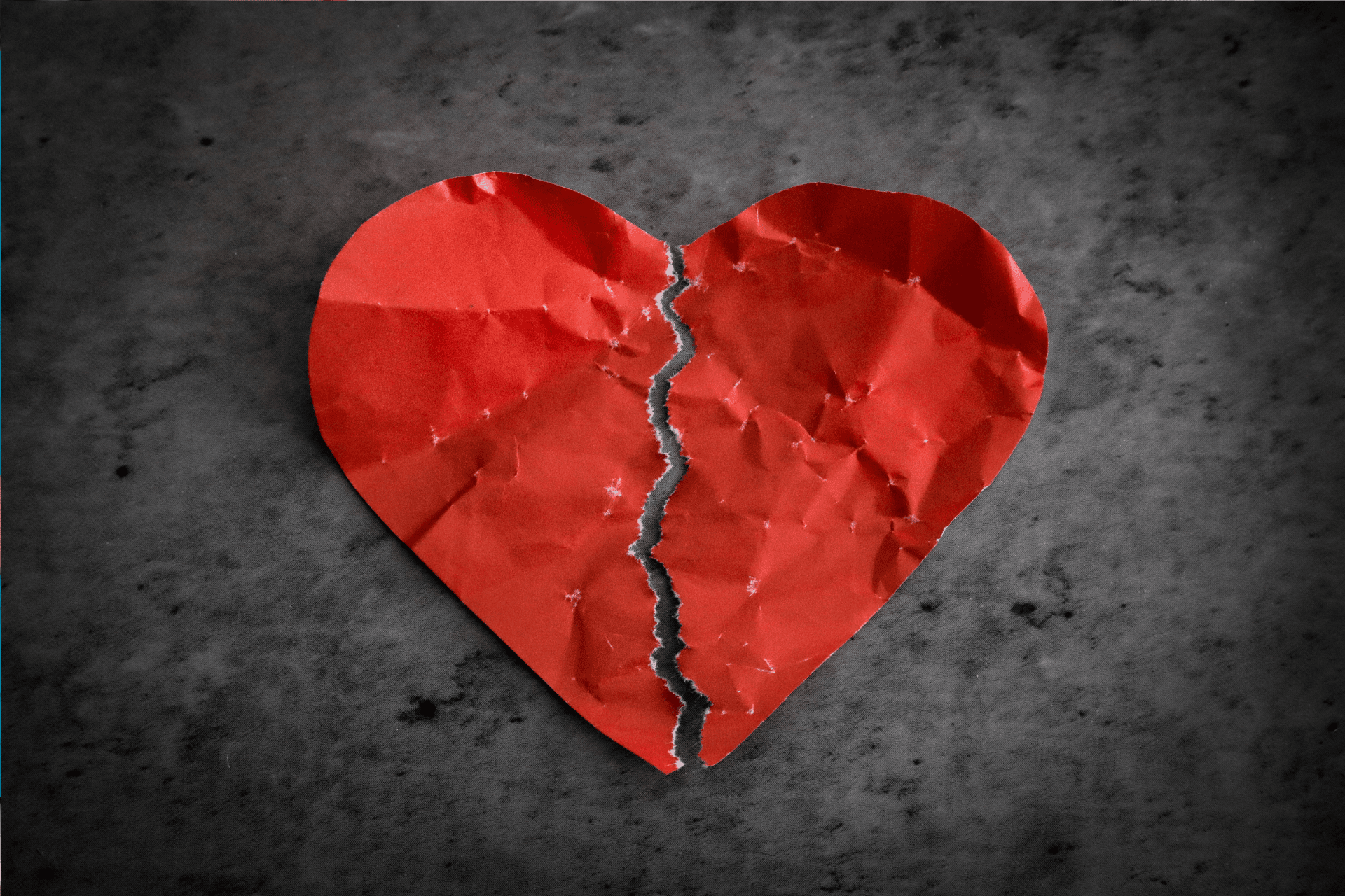 Crumpled red paper heart torn down the middle on a dark surface.