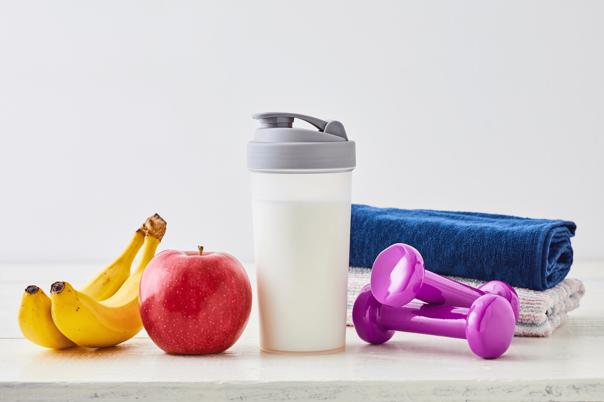 Healthy lifestyle setup with bananas, an apple, a protein shake, dumbbells, and towels.
