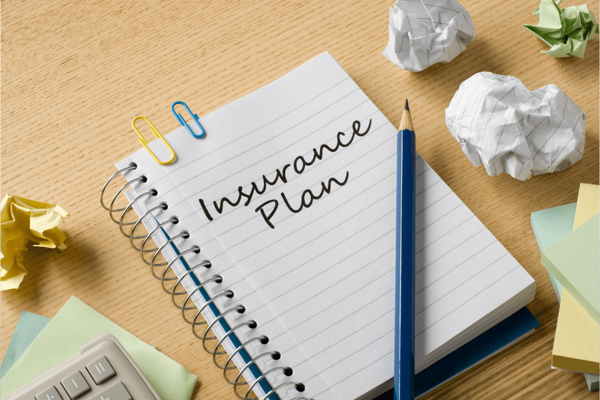 Notebook with the words ‘Insurance Plan’ written on it beside a pencil and crumpled paper