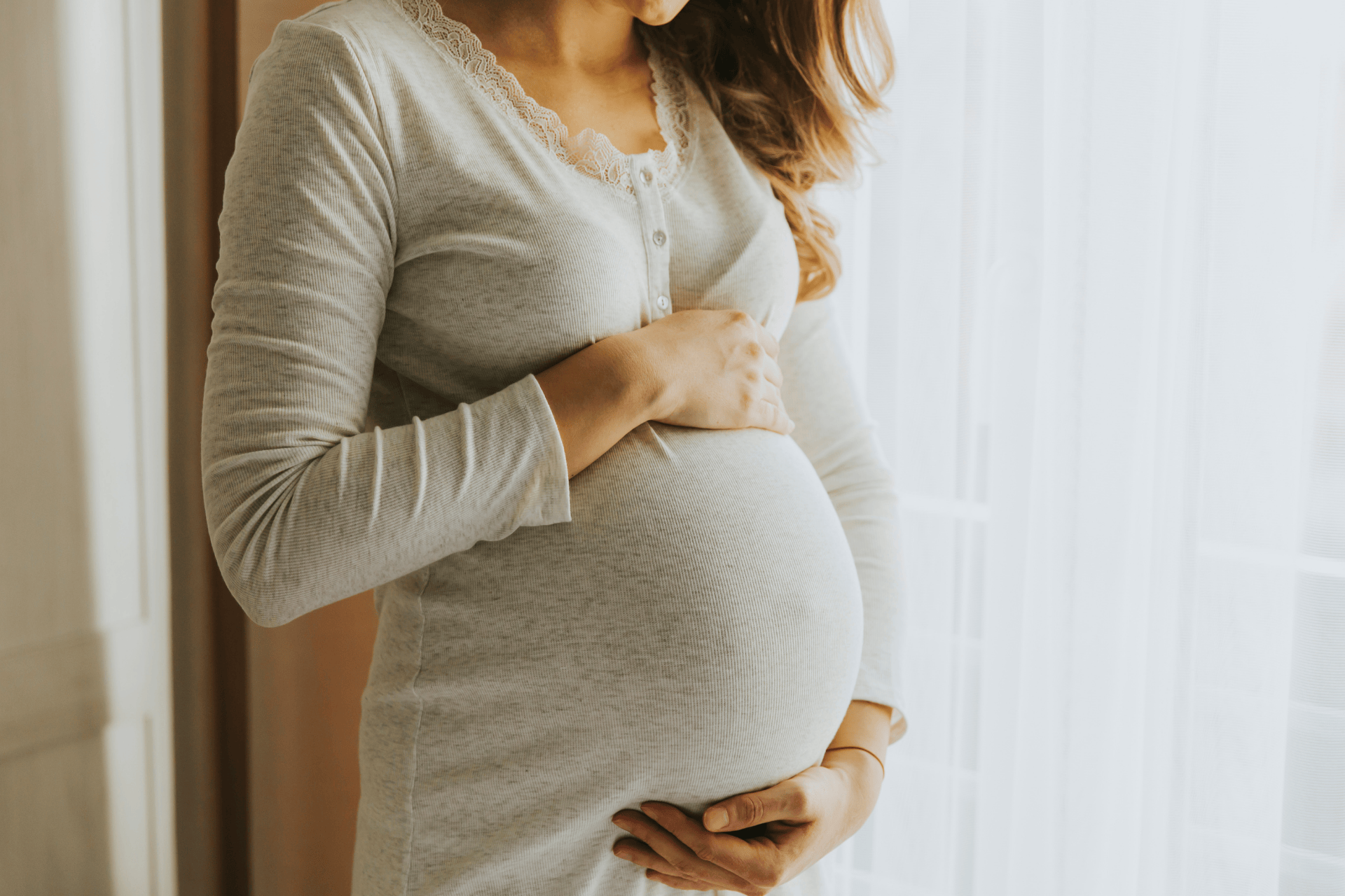 Pregnant woman gently holding her belly near a window with soft light.