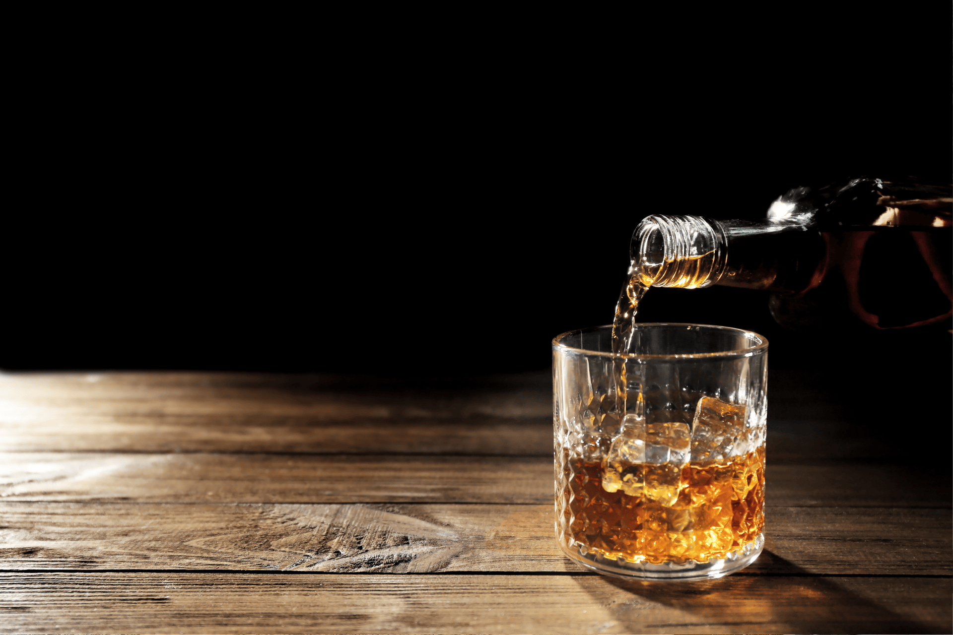 Whiskey being poured from a bottle into a glass with ice on a wooden table.