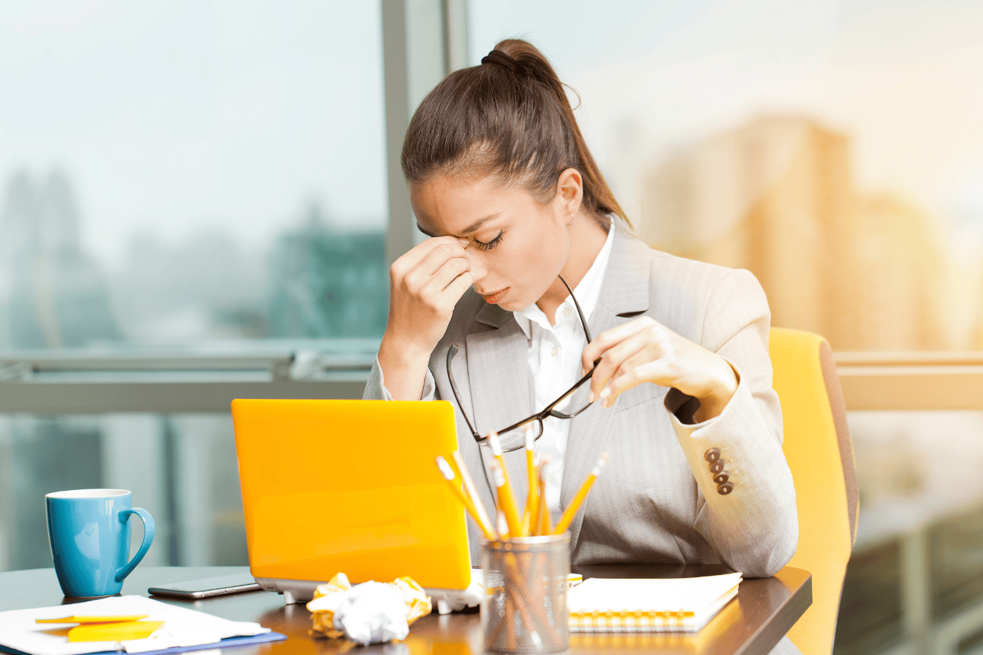 Stressed businesswoman sitting at desk, holding glasses and rubbing her eyes.