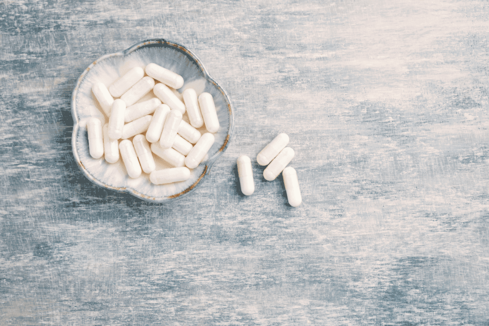 White capsules in a small decorative bowl with a few scattered on a textured surface.