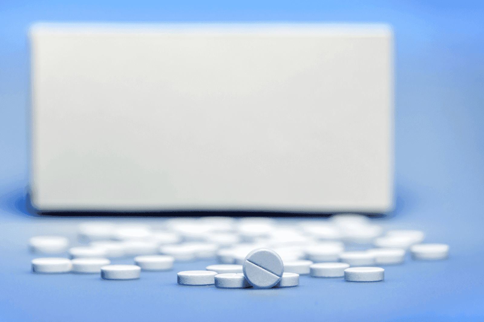 White tablets scattered on a blue surface in front of a plain white medicine box
