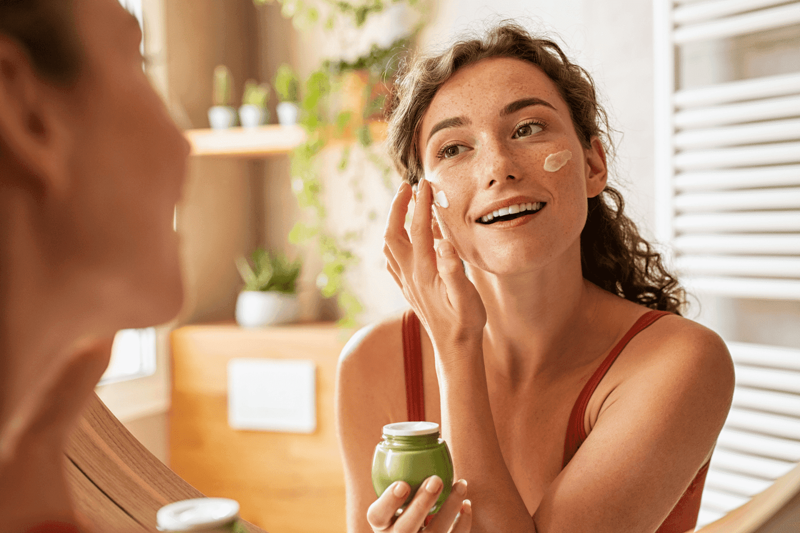 Woman applying face cream while smiling in a mirror.