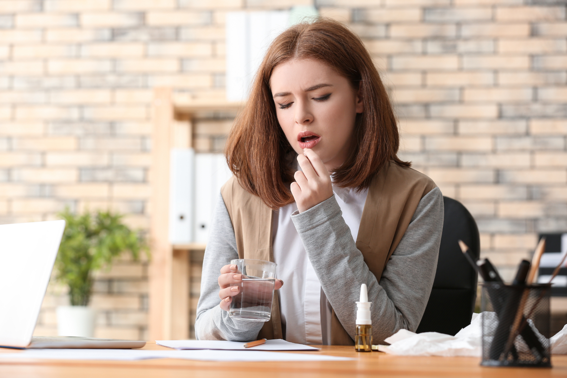 Woman at desk taking medication with a glass of water, appearing unwell indoors