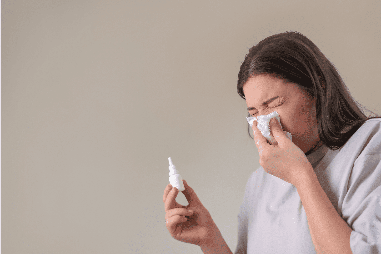 Woman blowing her nose with a tissue while holding a nasal spray.