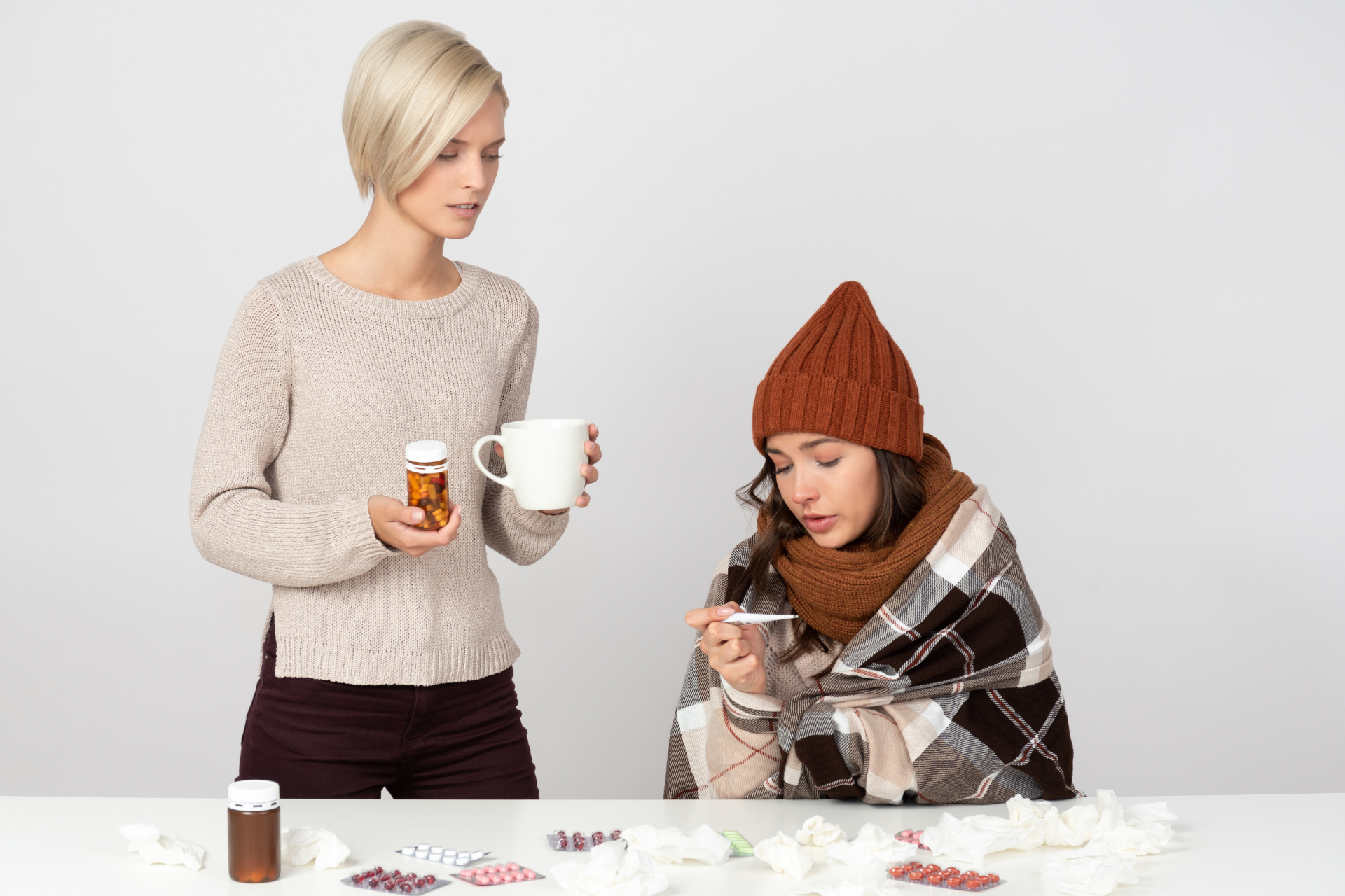 Woman caring for a sick friend wrapped in a blanket, offering medicine and a warm drink