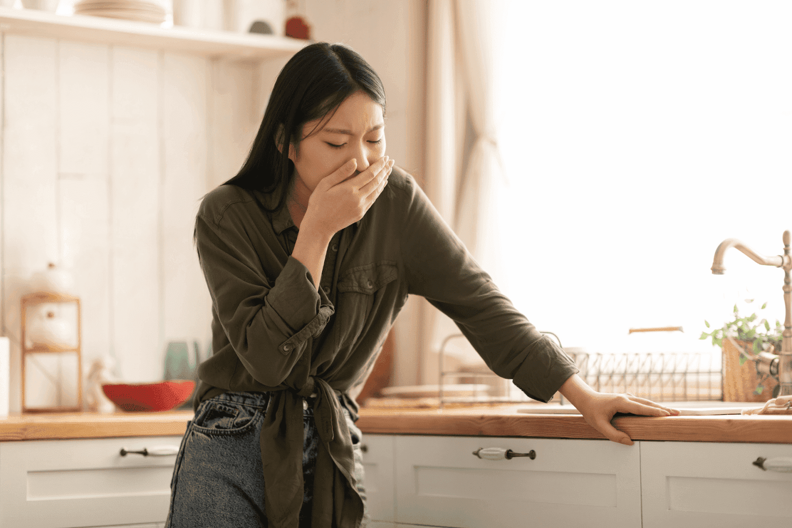 Woman covering her mouth and leaning on a kitchen counter, looking nauseous.