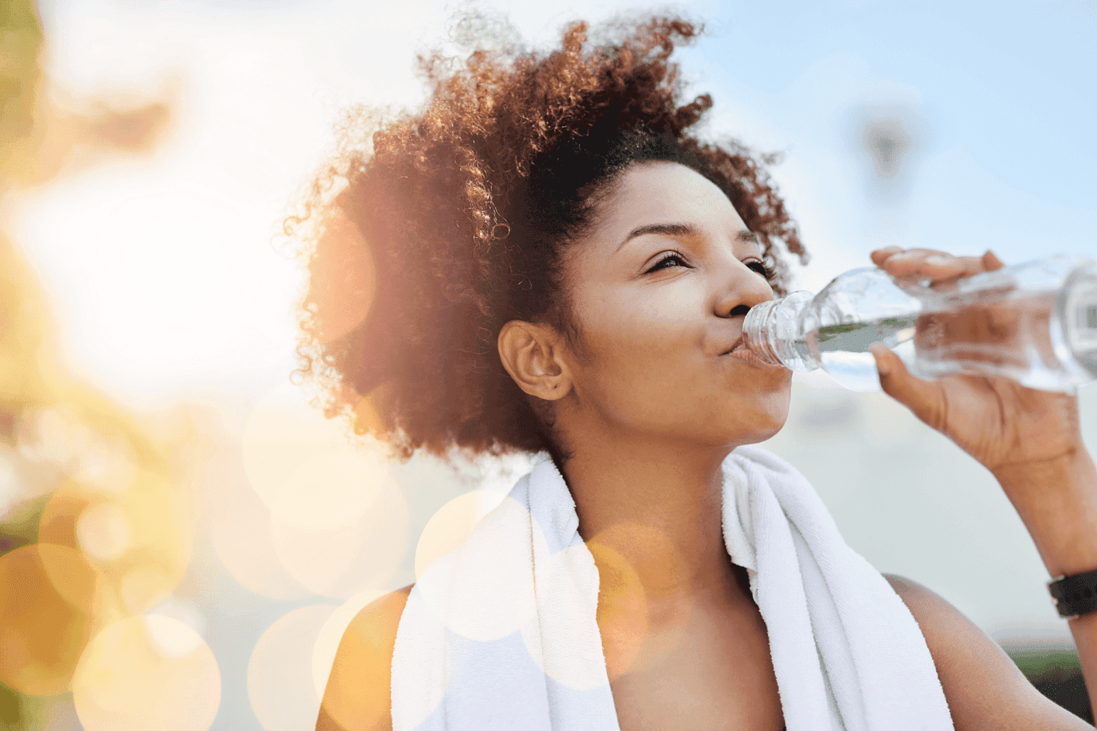 Woman drinking from a water bottle outdoors with sunlight in the background.