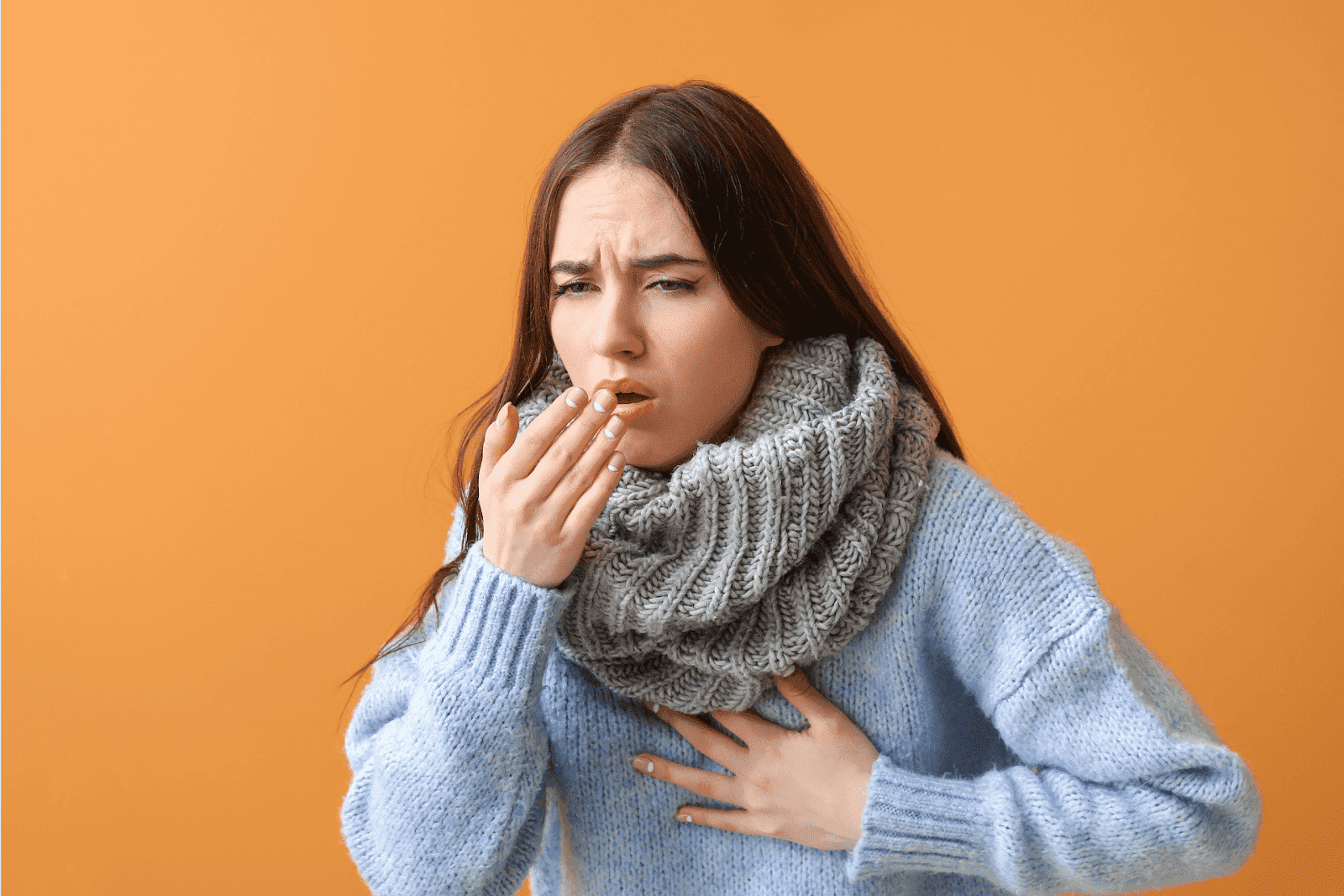 Woman in a blue sweater and scarf, coughing with her hand on her chest.