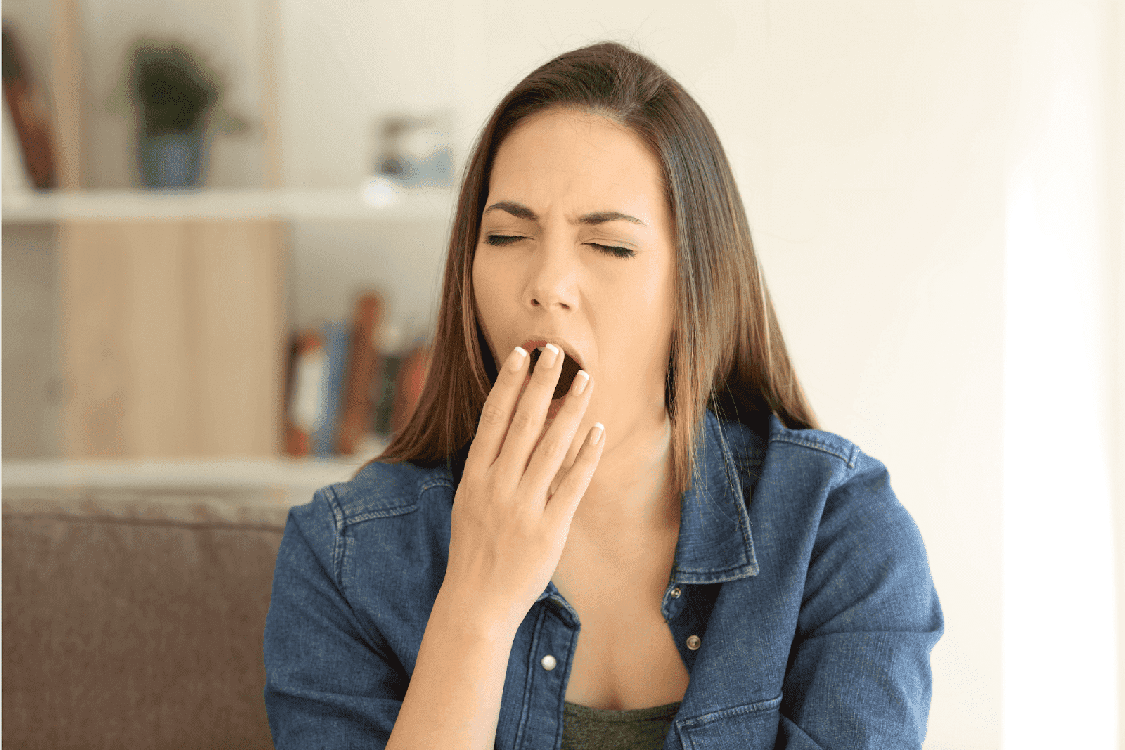 Woman in a denim shirt yawning with her hand covering her mouth.