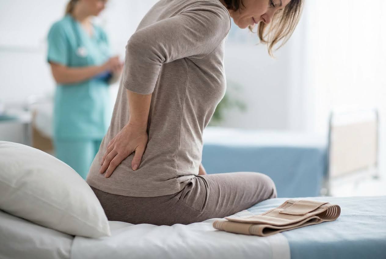 Woman in a hospital gown sitting on a bed, holding her lower back in pain, with a nurse in the background