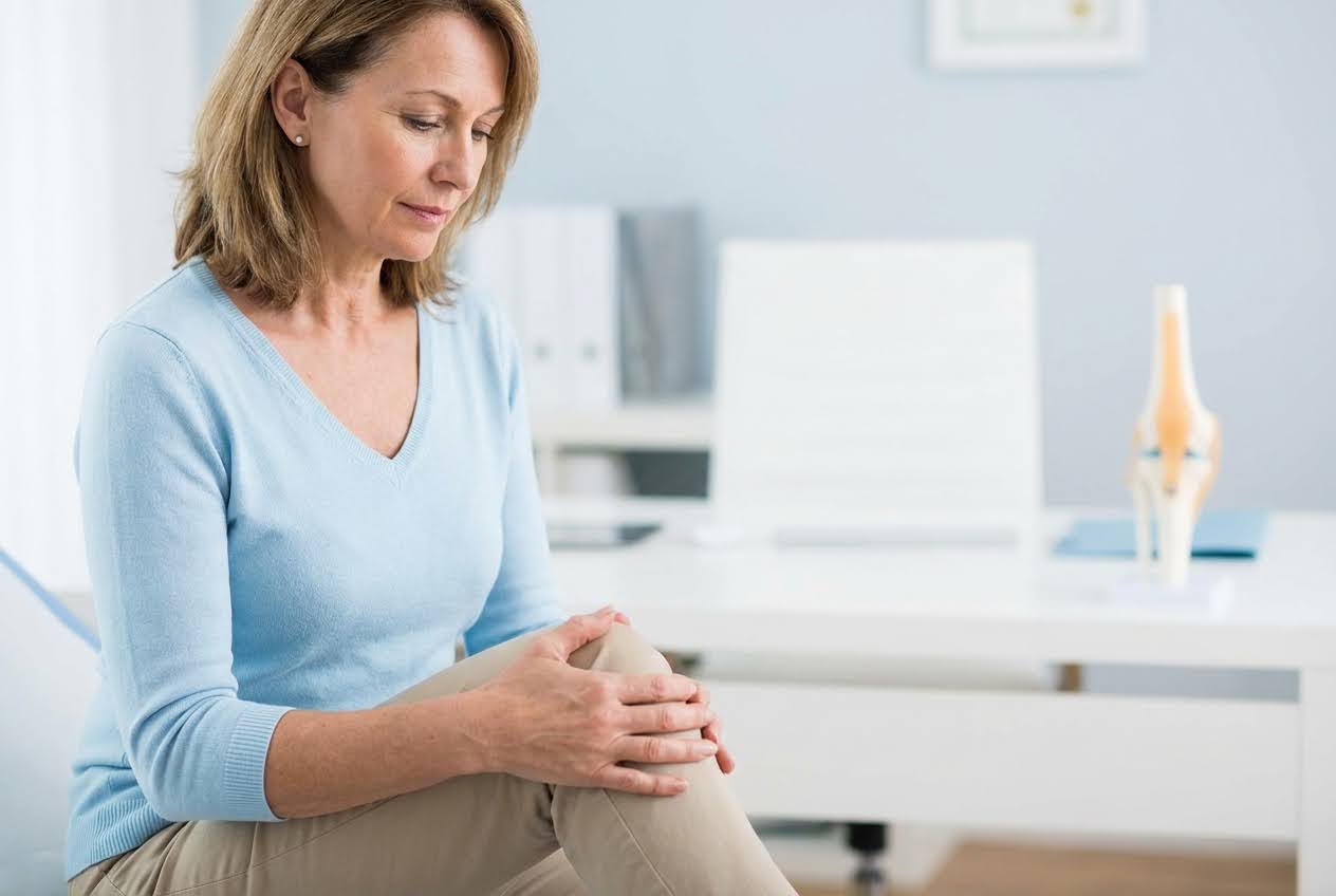 Woman in a light blue shirt holding her knee, with a knee joint model on a white desk in the background