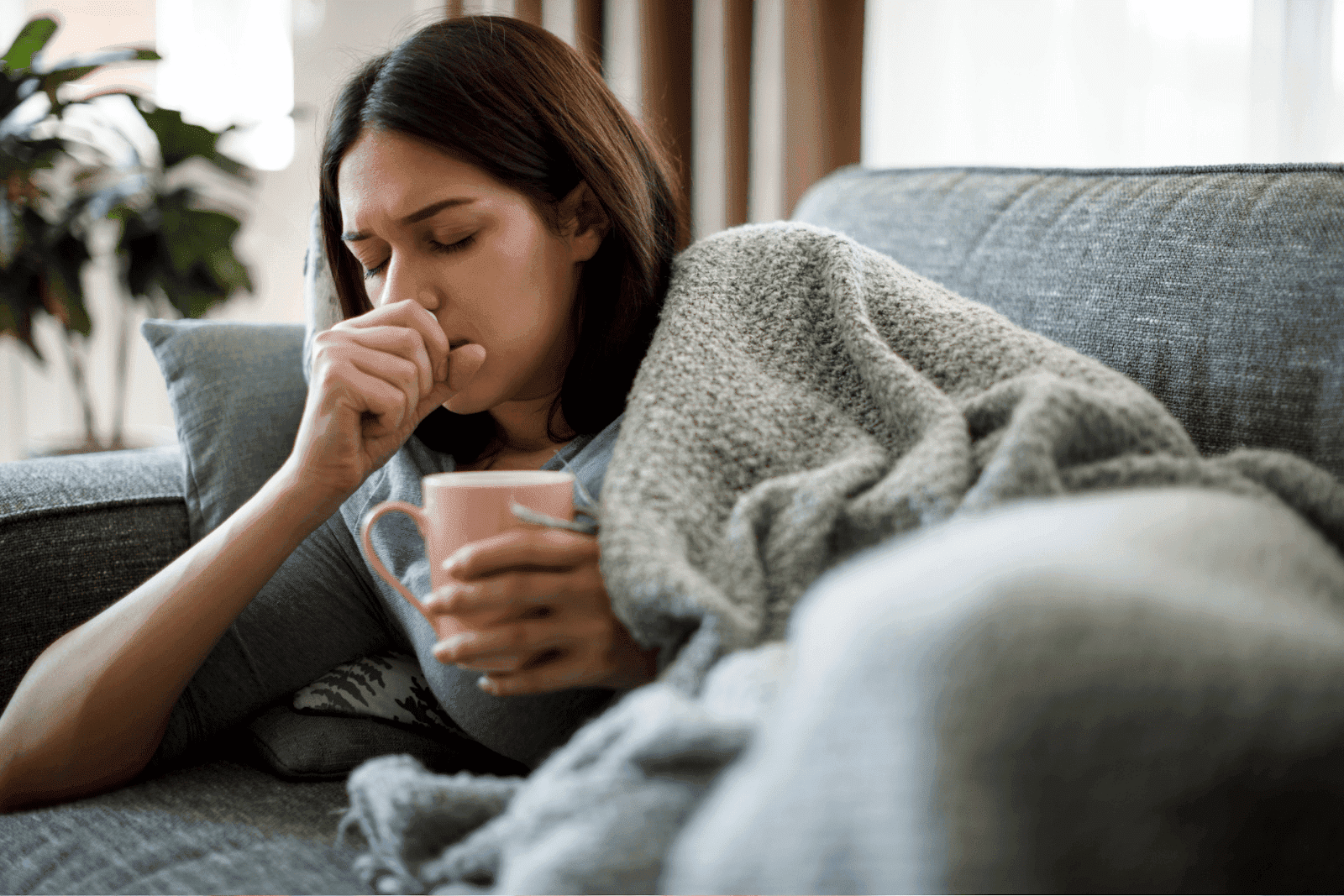 Woman lying on a couch under a blanket, coughing and holding a pink mug.